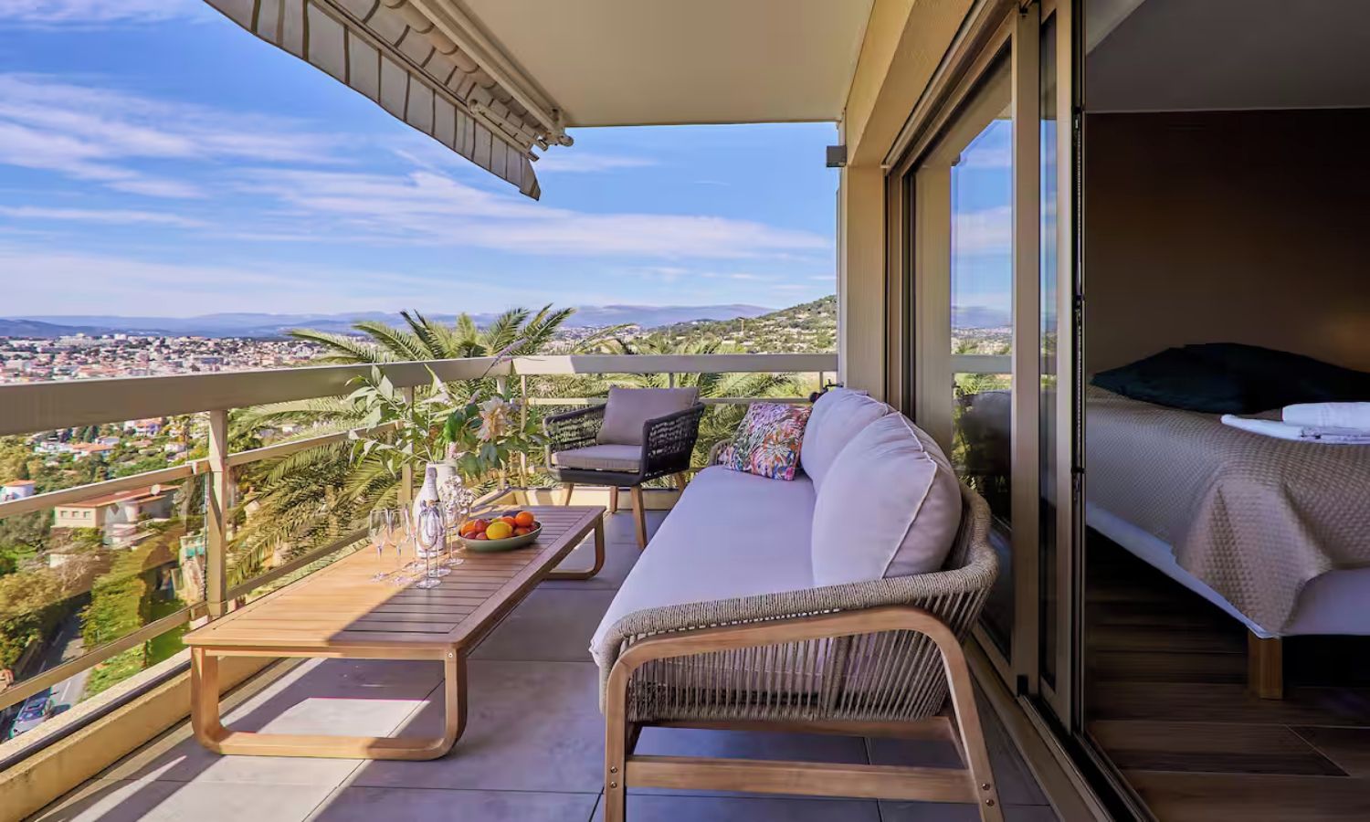 A balcony with a wicker sofa, a wooden table with a fruit bowl, and a view of the cityscape.
