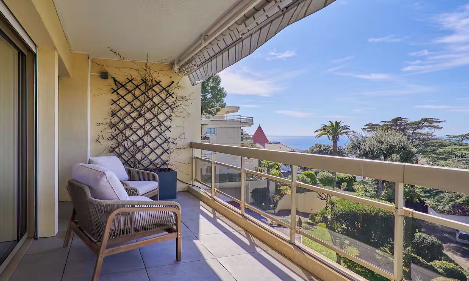Un balcon avec un fauteuil et des coussins en osier, un mur en treillis et une vue sur la mer et la verdure.