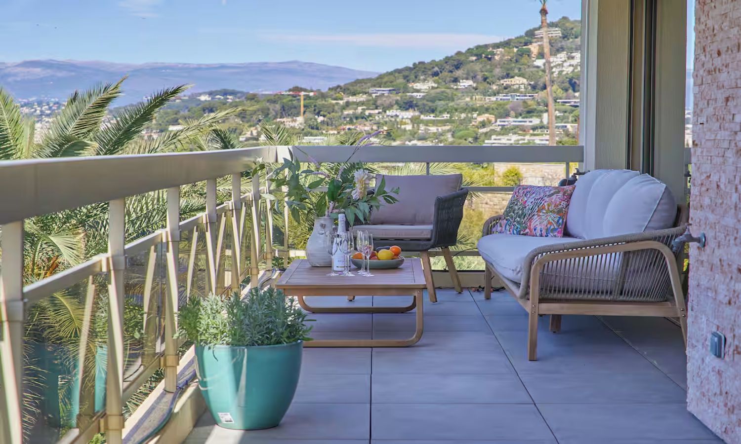 A balcony with a wicker sofa, a small table with a vase and fruit, and a potted plant.