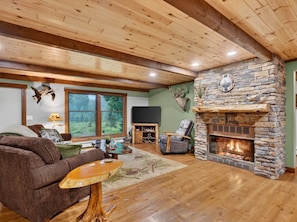 A cozy living room with a stone fireplace, wooden ceiling beams, and a large window overlooking greenery. - Escape to Buck Fever Lodge! An Exclusive Trophy Whitetail Ranch Experience! (Gladwin)