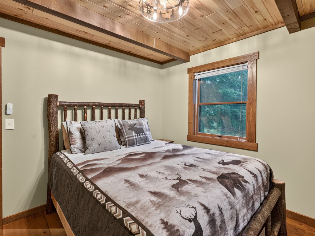 A bedroom with a wooden bed frame, a deer-themed bedspread, a window with a view of trees, and a wooden ceiling.