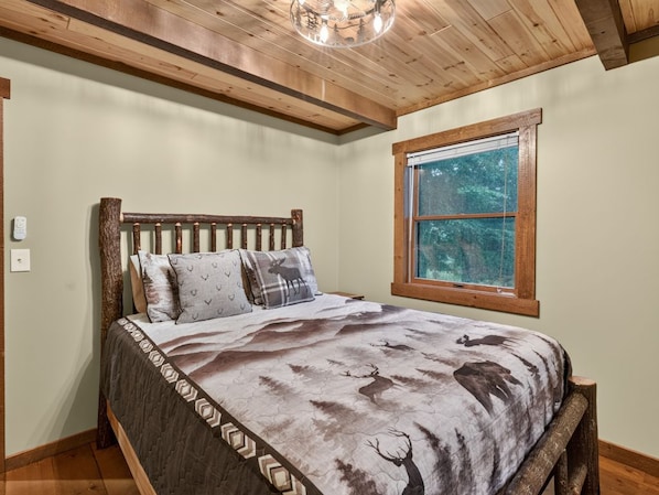 A bedroom with a wooden bed frame, a deer-themed bedspread, a window with a view of trees, and a wooden ceiling.