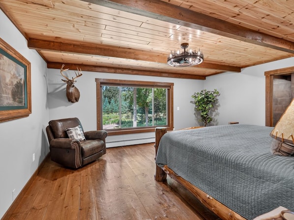 A bedroom with a wooden ceiling, a large window, a bed, a chair, and a wall-mounted deer head.