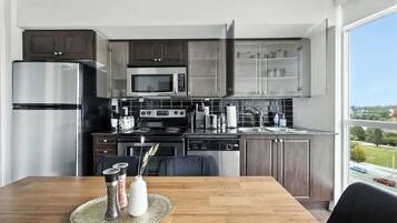 A modern kitchen with stainless steel appliances, a dining area with a wooden table, and a view of the outdoors through a large window.