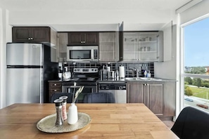 A modern kitchen with stainless steel appliances, a dining area with a wooden table, and a view of the outdoors through a large window.