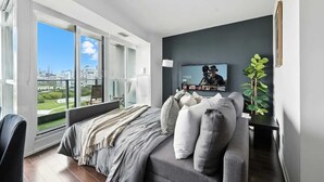 Quarto moderno com uma janela grande, cama com roupa de cama cinzenta, televisão na parede e uma planta.