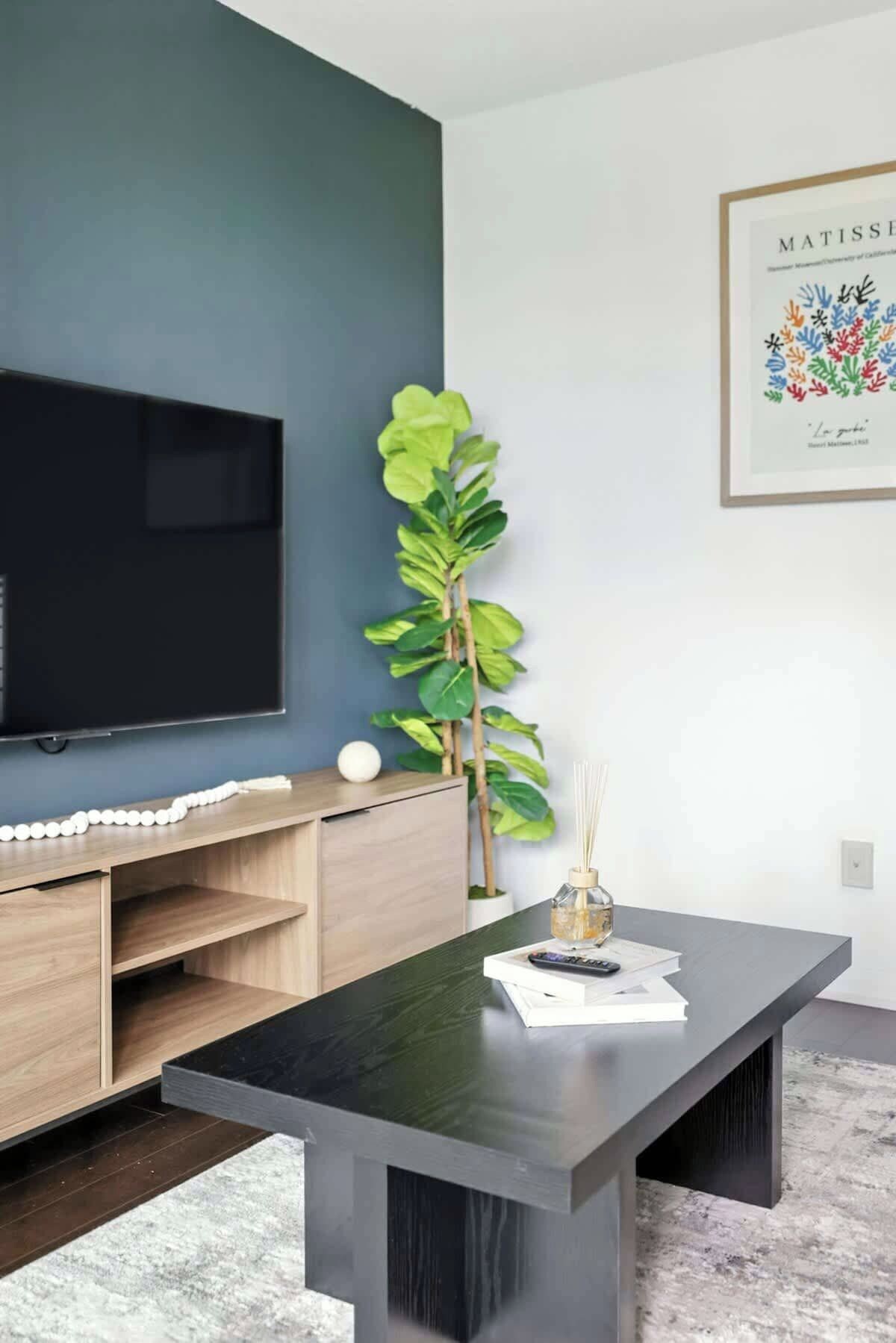 A modern living room with a flat-screen TV, a wooden entertainment unit, a large green plant, a dark coffee table, and a framed artwork on the wall.