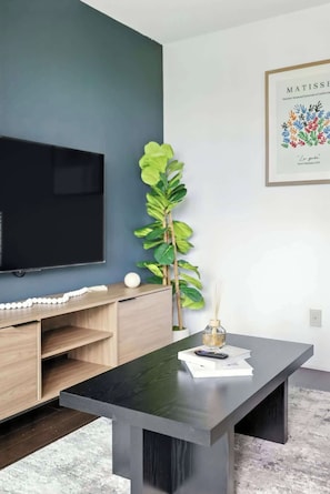 A modern living room with a flat-screen TV, a wooden entertainment unit, a large green plant, a dark coffee table, and a framed artwork on the wall.