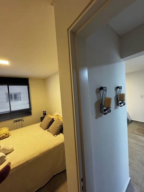 A bedroom with a bed, pillows, a bedside table, a window with blinds, and a door with wall-mounted light fixtures. - Modern, comfortable apartment in Salta with an excellent location — VRTEAM (Salta)