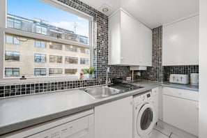Eine moderne Küche mit einer integrierten Waschmaschine, einem Spülbecken und einem Fenster mit Blick auf ein Wohngebäude.