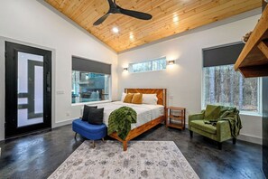 A bedroom with a bed, a blue chair, a green chair, a wooden ceiling fan, and a window with a view of trees. - Sauna, Hot Tub, Fire Pit, Pets, EV Plug, Secluded (Broken Bow)