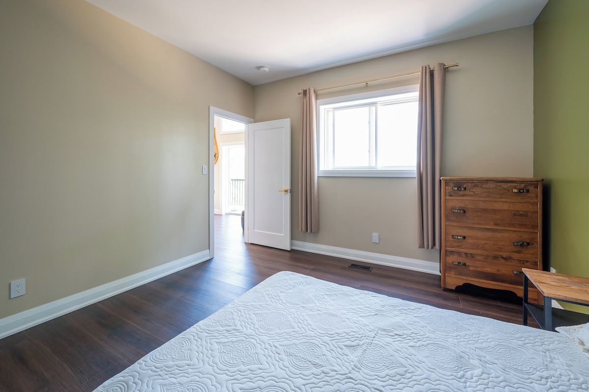 A bedroom with a large bed, a dresser, a window with curtains, and a door leading to another room.