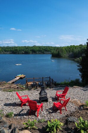 A lakeside deck with red chairs and a fire pit, overlooking a calm lake with boats.