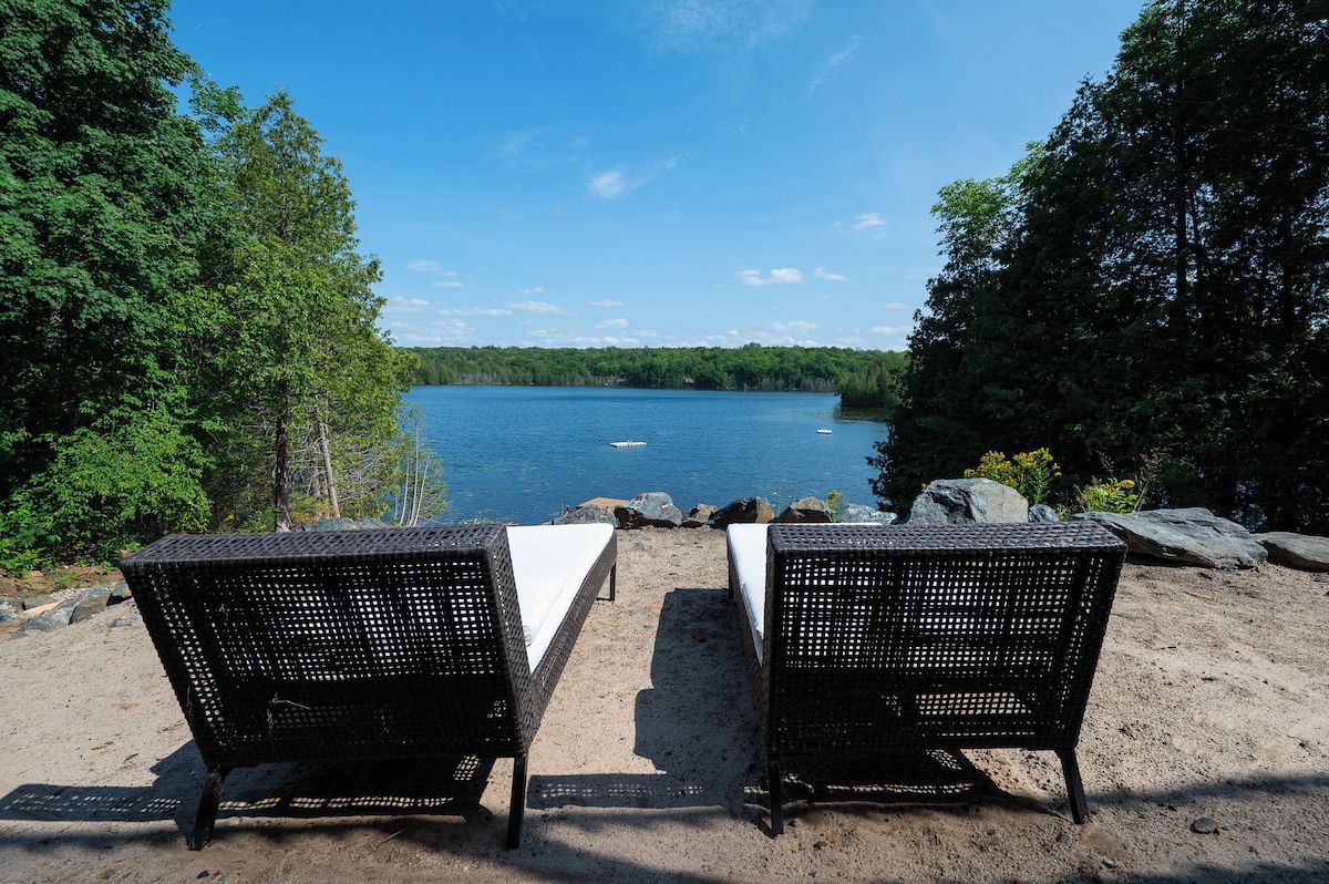 Two wicker chairs with white cushions on a sandy beach, overlooking a calm lake with boats, surrounded by dense green trees.