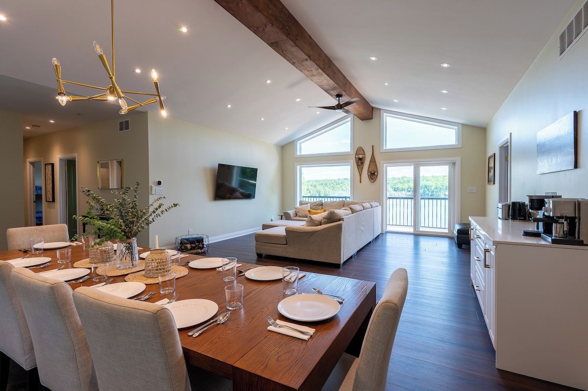 A spacious dining area with a wooden table set for a meal, a sofa in the background, and a kitchen area with a coffee machine.