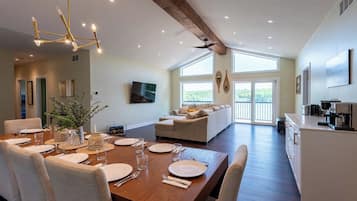 A spacious dining area with a wooden table set for a meal, a sofa in the background, and a kitchen area with a coffee machine.