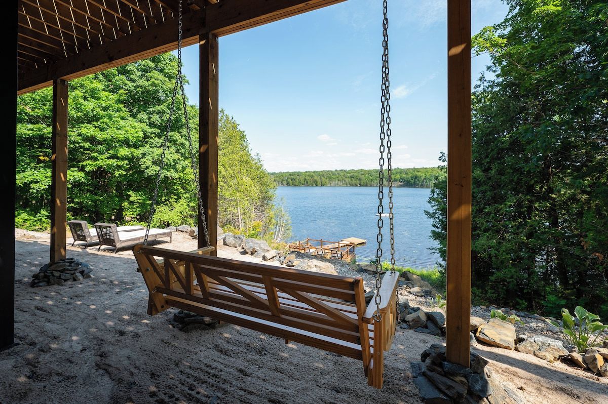 Un banco de madera para columpios bajo un porche con vista a un lago y bosque.