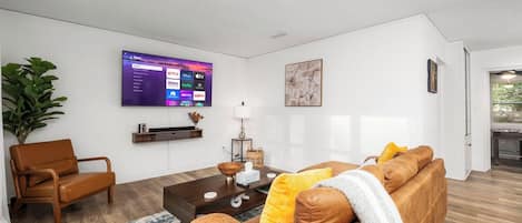 A modern living room with a flat-screen TV mounted on the wall, a brown leather sofa, a wooden coffee table, and a potted plant.