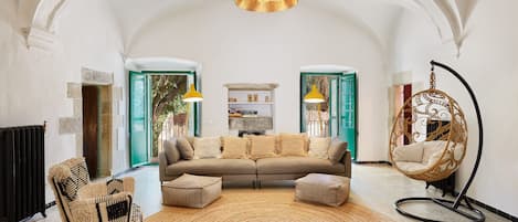En rummelig stue med et hvælvet loft, en brun sofa med puder, en rund vævet måtte, en hængestol og en radiator.
