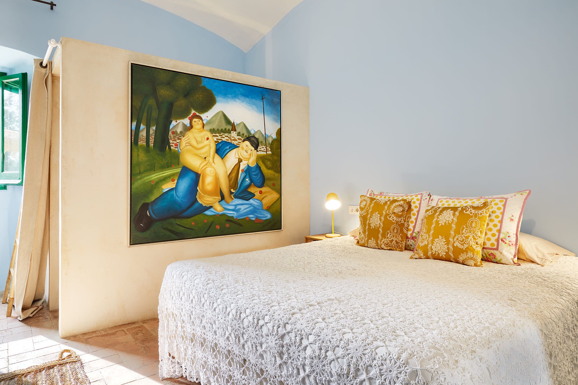 A bedroom with a large painting on the wall, a bed with a white lace cover, and patterned pillows.