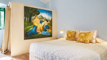 A bedroom with a large painting on the wall, a bed with a white lace cover, and patterned pillows.