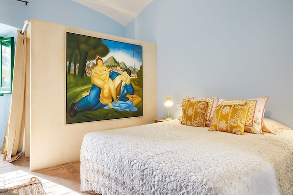 A bedroom with a large painting on the wall, a bed with a white lace cover, and patterned pillows.