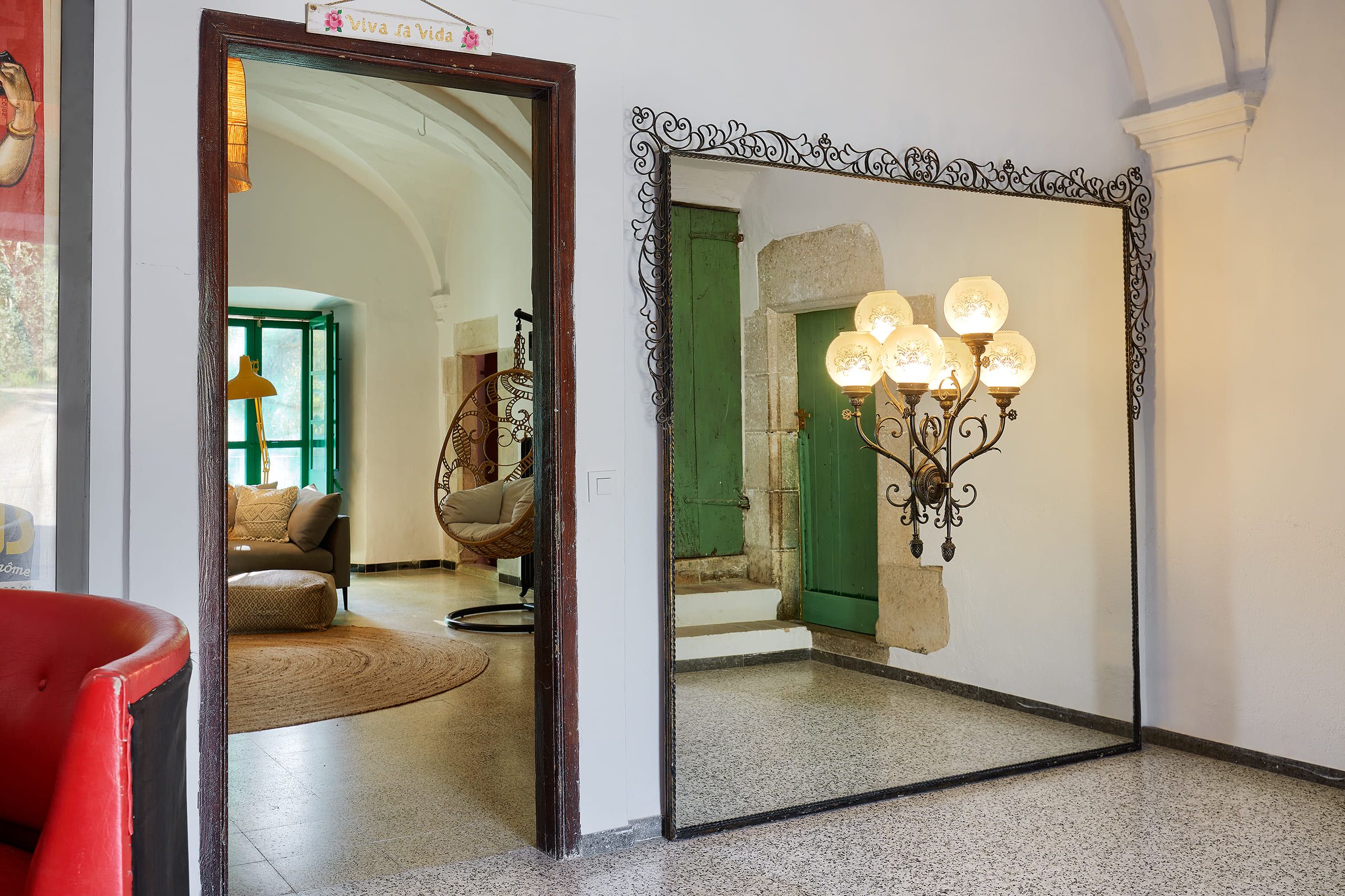 Ein Zimmer mit einem großen Spiegel, der eine grüne Tür, einen Korbstuhl und eine Stehlampe reflektiert.