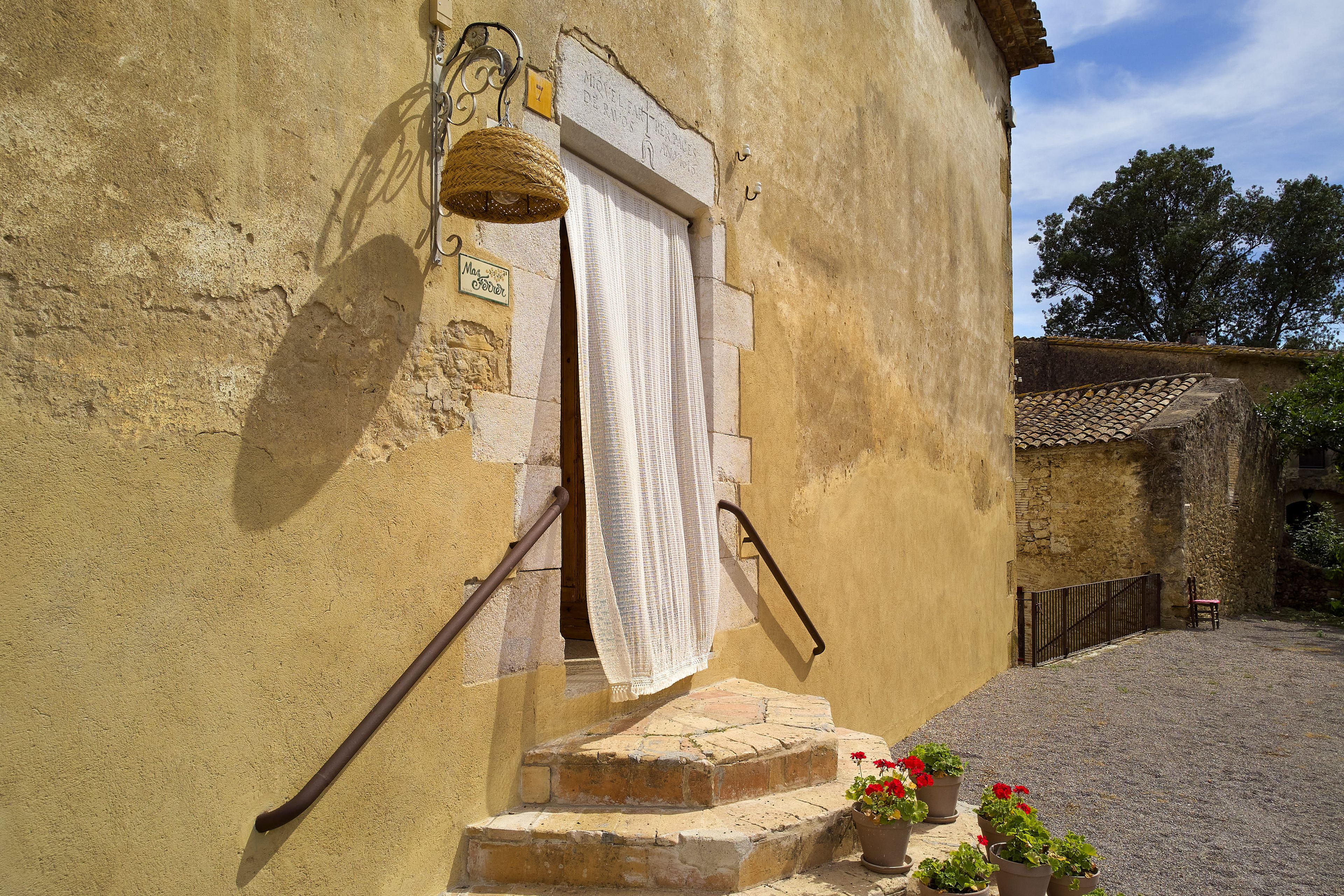 A rustic building with a stone staircase leading to a door with a white curtain. There are potted plants on the steps and a metal gate in the background.