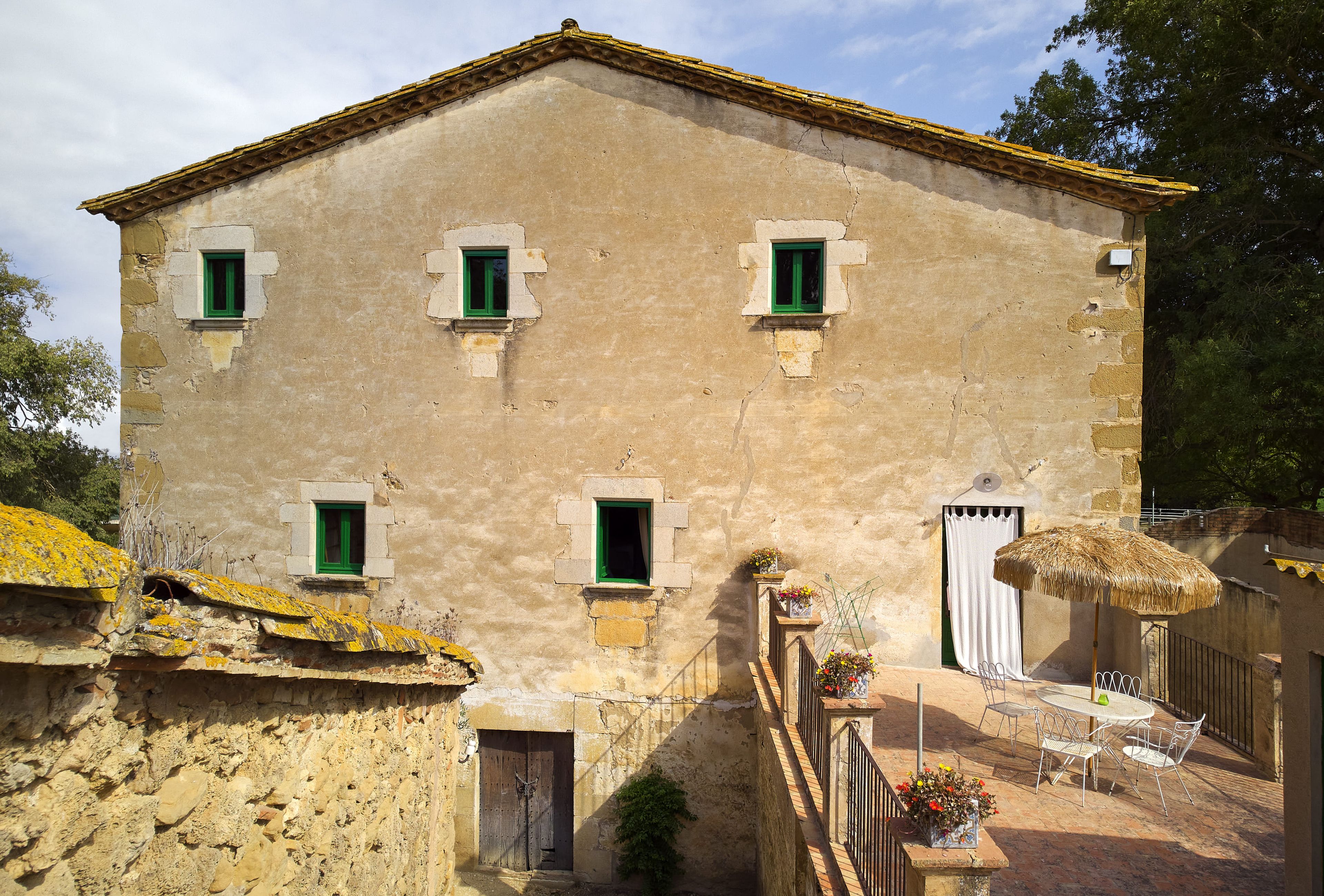 A two-story building with a stone facade, multiple windows with shutters, a wooden door, and a small outdoor seating area with a table and chairs.