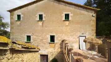 A two-story building with a stone facade, multiple windows with shutters, a wooden door, and a small outdoor seating area with a table and chairs.