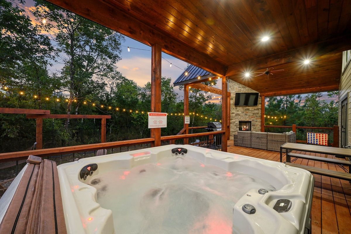A hot tub on a wooden deck with a stone fireplace and outdoor seating.