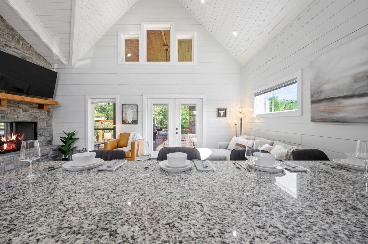 A spacious living area with a high ceiling, a fireplace, a dining table set for four, and a comfortable seating arrangement.