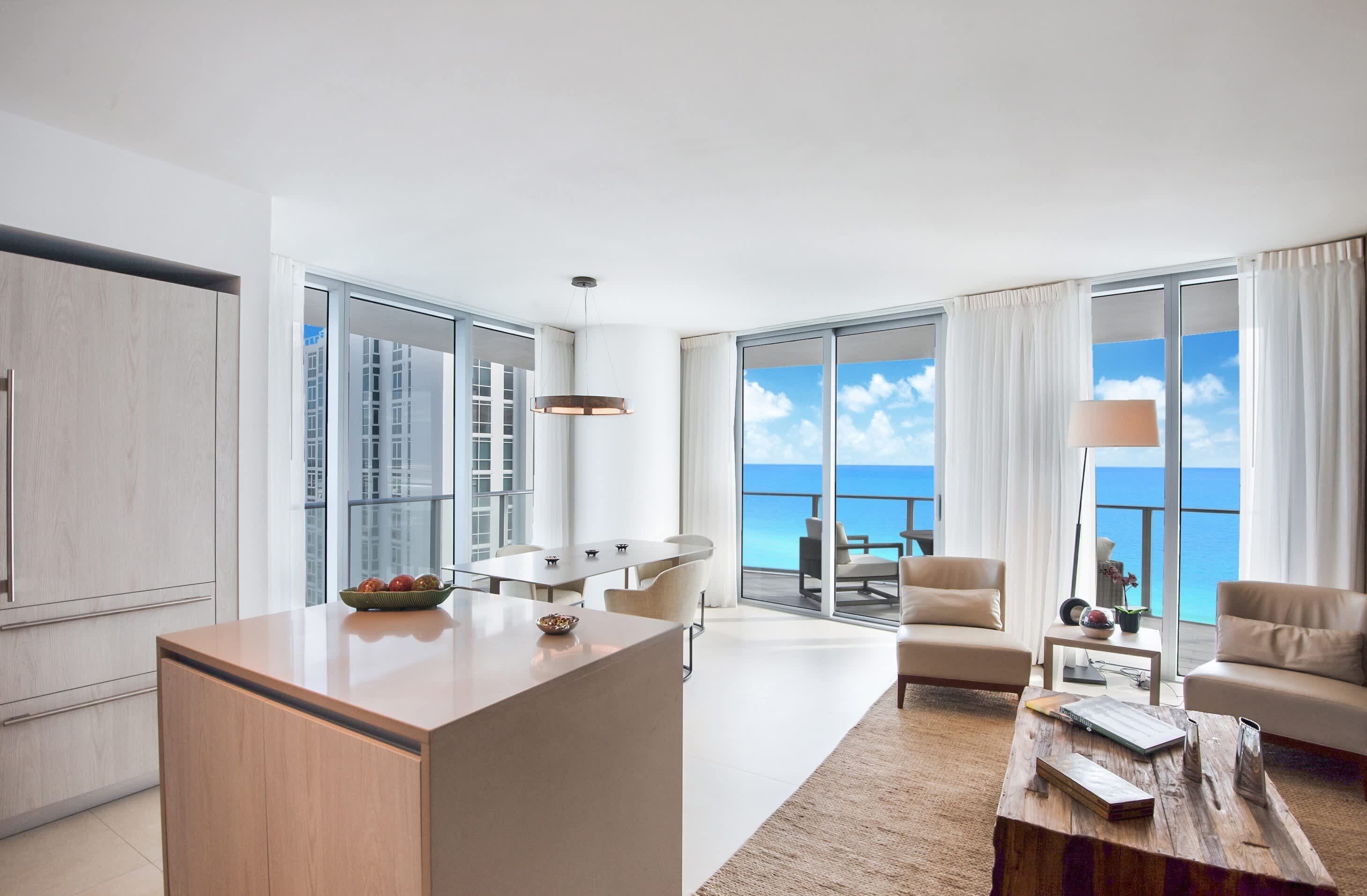 A modern hotel room with a large balcony, a dining area, and a seating space with a view of the ocean.