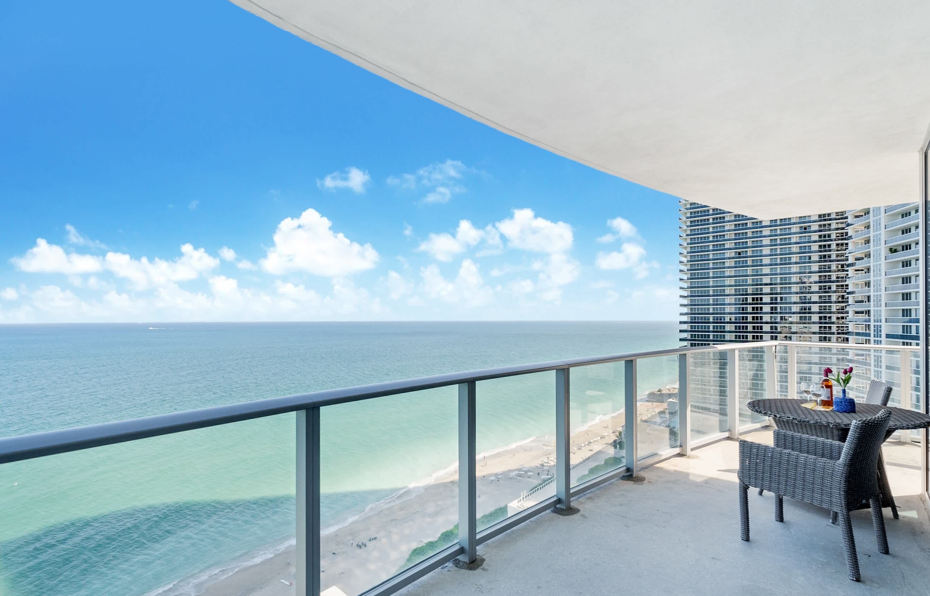 A balcony with a view of the ocean and a beach, a dining table with chairs, and a modern building in the background.
