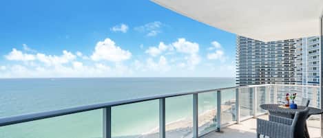 A balcony with a view of the ocean and a beach, a dining table with chairs, and a modern building in the background.