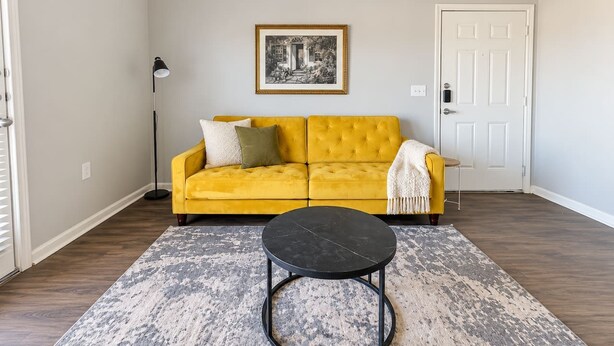 Una sala de estar con un sofá amarillo, una mesa de centro redonda negra y una alfombra estampada.