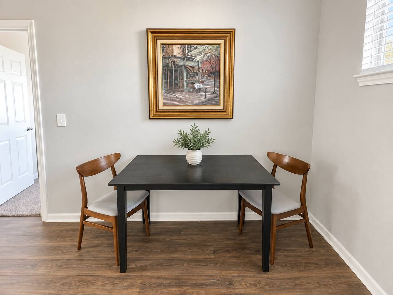 A dining table with two chairs, a framed painting above it, and a small plant on the table.