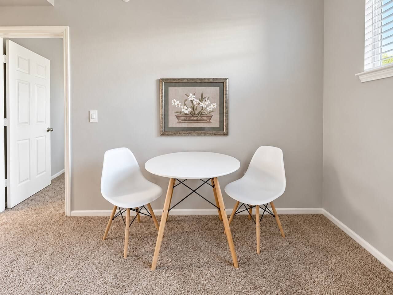 Een kamer met een witte deur, een lijst met een afbeelding aan de muur, en een ronde tafel met twee witte stoelen.