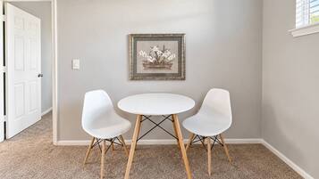 Een kamer met een witte deur, een lijst met een afbeelding aan de muur, en een ronde tafel met twee witte stoelen.