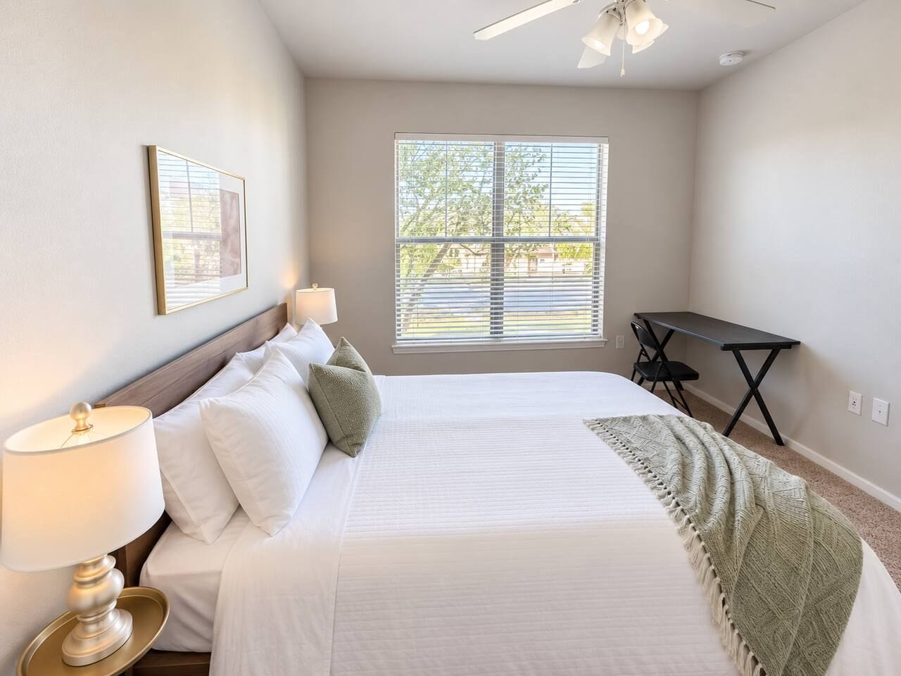 A bedroom with a bed, bedside lamp, window with blinds, and a desk with a chair.