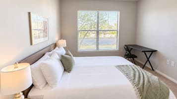 A bedroom with a bed, bedside lamp, window with blinds, and a desk with a chair.