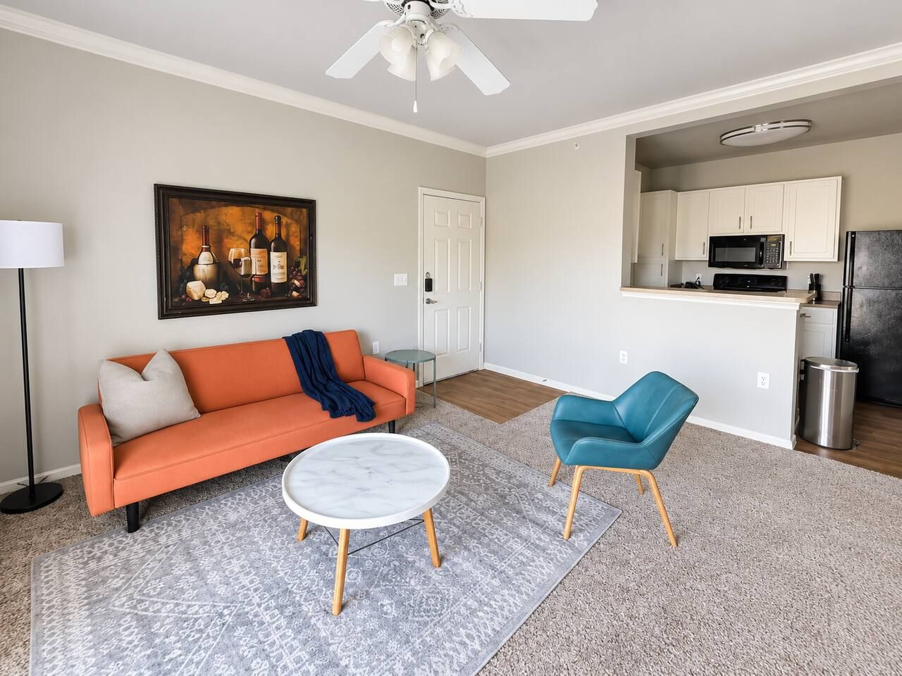 A living room with a sofa, a coffee table, a blue chair, a painting, and a kitchen area with cabinets and appliances.