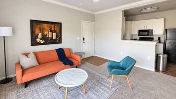 A living room with a sofa, a coffee table, a blue chair, a painting, and a kitchen area with cabinets and appliances.