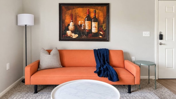 Ett vardagsrum med en orange soffa, ett runt, vitt soffbord, en målning som föreställer vinflaskor, en golvlampa och en dörr.