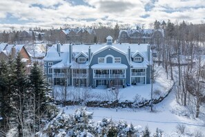 A large, multi-story house with a snow-covered roof and multiple chimneys, surrounded by trees and other houses. - The Sanctuary at Les Manoirs (Mont Tremblant)
