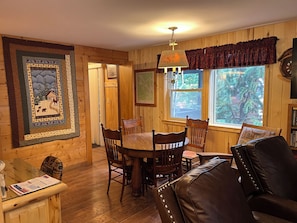 L'intérieur d'un chalet accueillant, avec une table à manger, des chaises, un canapé en cuir, un lustre et une grande fenêtre donnant sur la forêt.