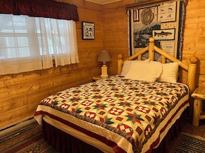 Une pièce de cabane en bois comprenant un lit, une fenêtre avec des rideaux, un mur orné d’une enseigne sur le thème des poissons et une petite table avec une lampe.