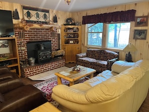 A cozy living room with a brick fireplace, a sofa, a coffee table, and a television. - Three Pines Cabin - Cozy & Comfortable 3 Bedroom (South Fork)