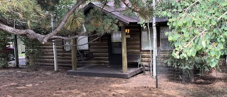 A log cabin with a porch, surrounded by trees and a fenced area.