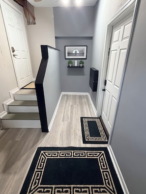 A hallway with a staircase, a framed picture, a small shelf with plants, and a black rug with a geometric pattern.
